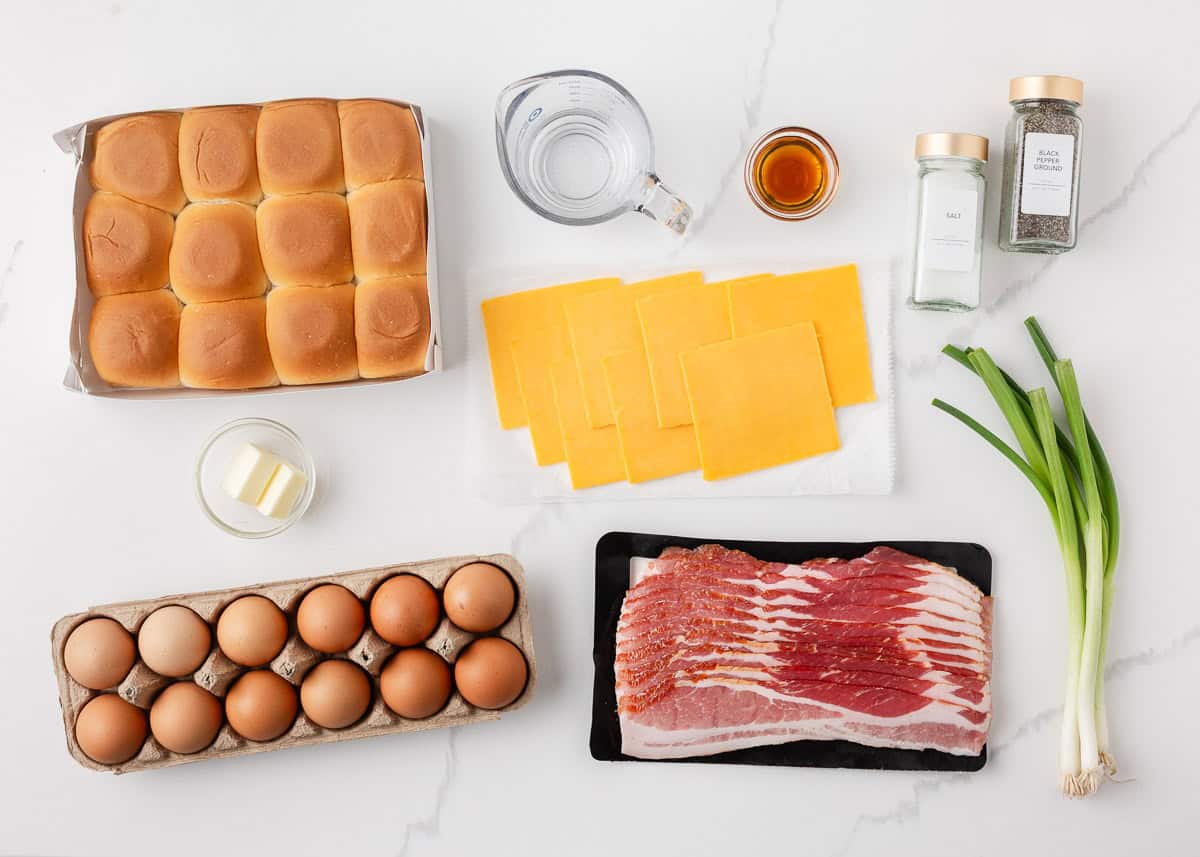Wow your breakfast or brunch crowd with these crispy golden buns filled with bacon, eggs, and cheddar cheese! Baked as one, then pulled apart, these Hawaiian roll breakfast sliders are easy to make and even easier to serve! Flat lay of Hawaiian roll breakfast slider ingredients including a pack of Hawaiian rolls, eggs, raw bacon strips, sliced cheddar cheese, butter, maple syrup, water, salt, black pepper, and green onions on a white marble surface.