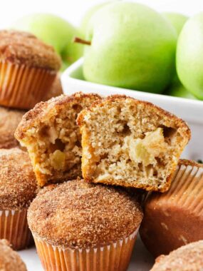 Close-up of a cinnamon apple muffin torn open to reveal a moist interior with diced apple chunks, nestled among sugar-coated muffins and fresh green apples in the background.