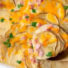 A gold spoon scooping cheesy scalloped potatoes and ham from a baking dish showing the creamy layers.
