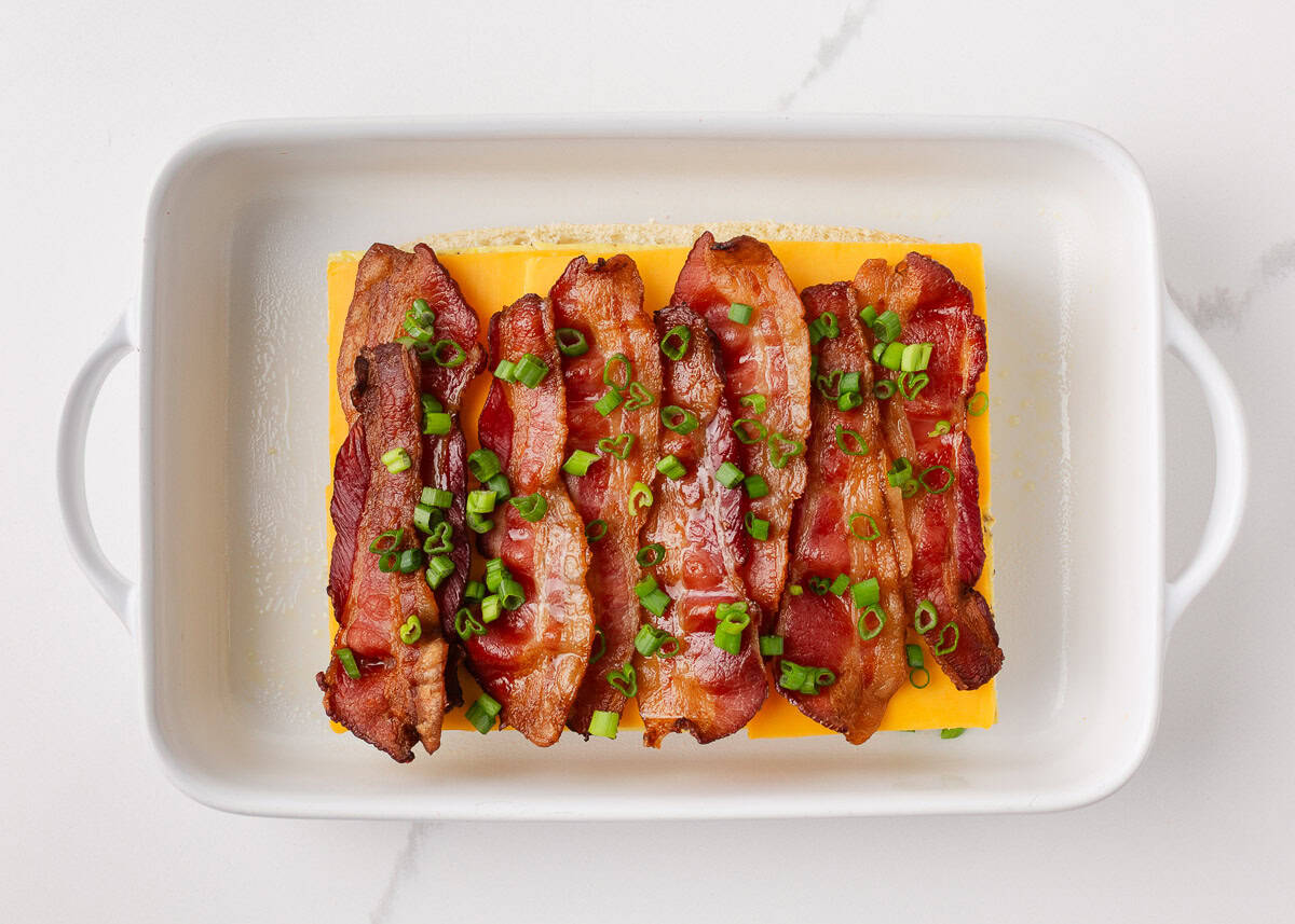 Overhead view of cooked bacon strips and sliced green onions layered on top of cheddar cheese and a baked sheet egg inside a white baking dish for breakfast sliders.