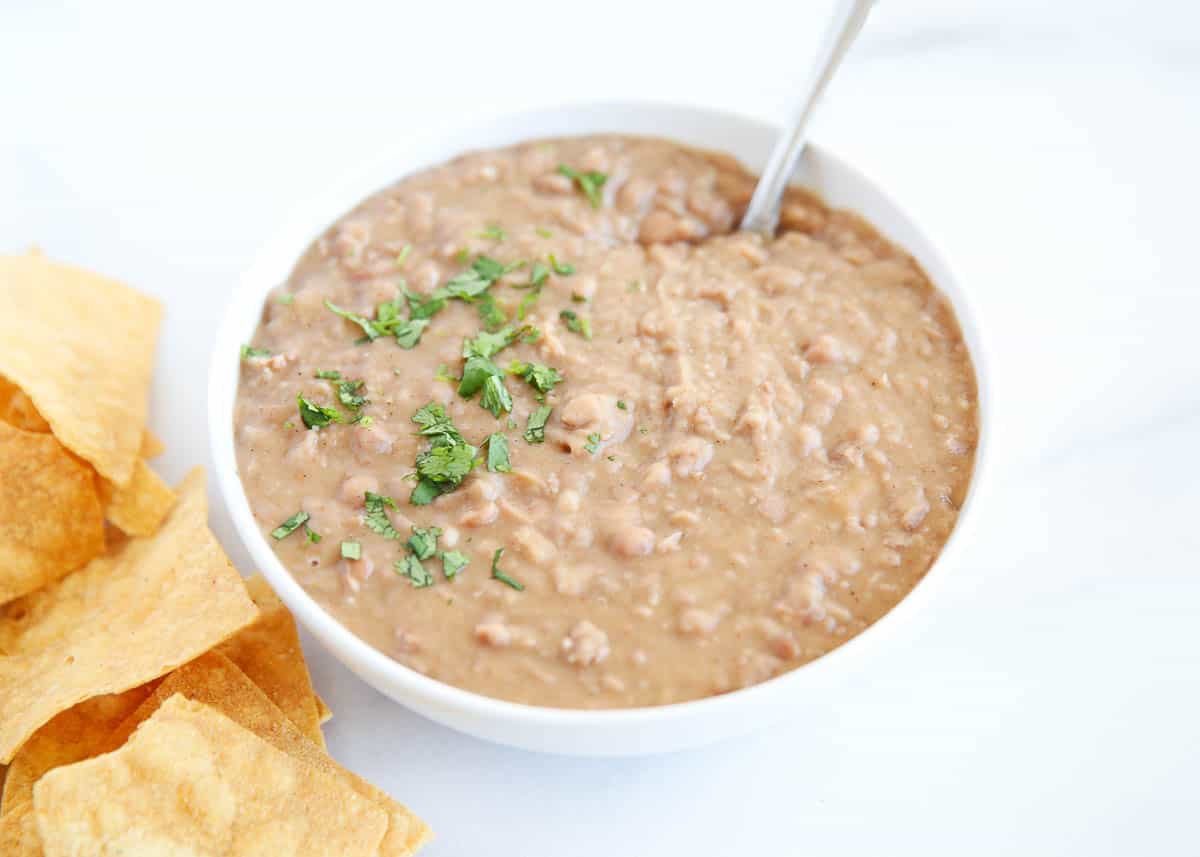 Easy refried beans in a white bowl with serving spoon and tortilla chips on the side.