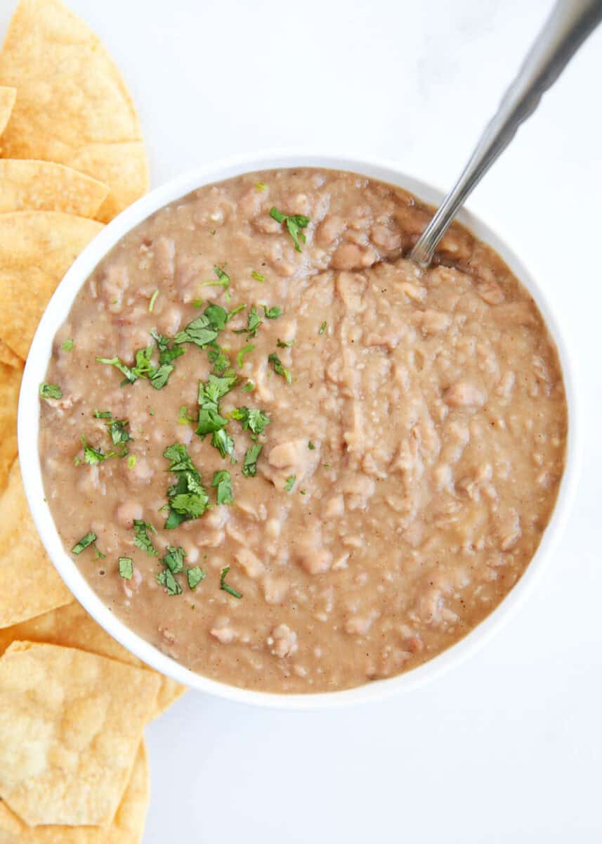 Refried beans in a white bowl with serving spoon and cilantro garnish.