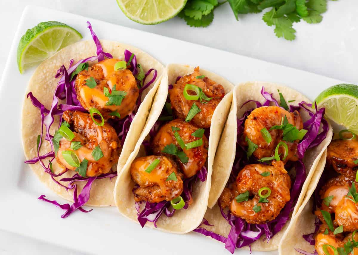 Throw away the takeout menu and enjoy making these bang bang shrimp tacos at home! Crispy fried shrimp are tossed in a creamy sweet and spicy sauce and then piled into corn tortillas and topped with crunchy cabbage. Dinner is ready in under 30 minutes! Bang bang shrimp tacos on a white plate.