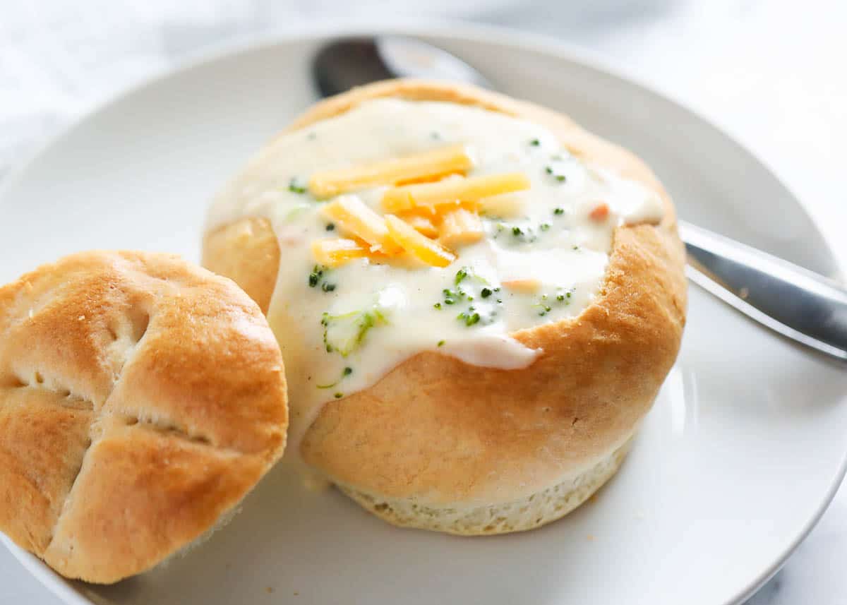 A bread bowl filled with broccoli cheddar cheese on a white plate with a silver spoon. 