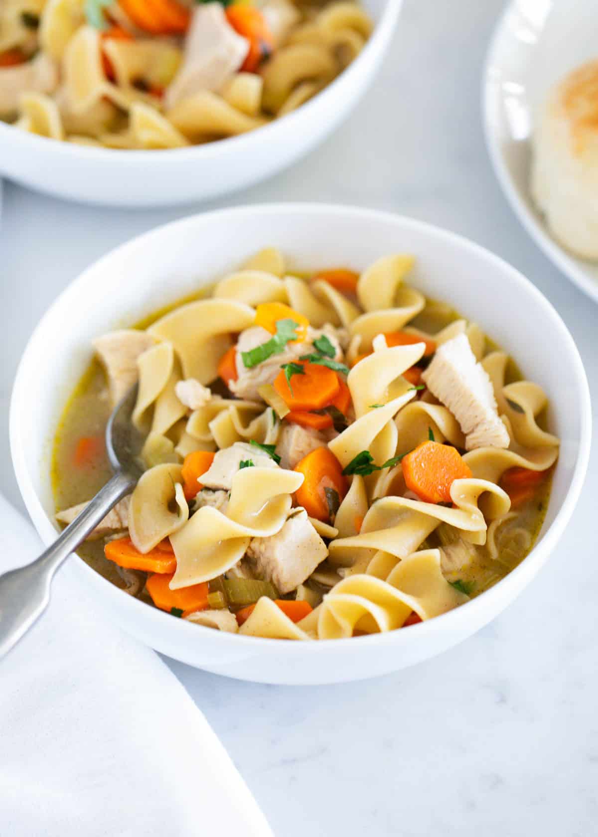 Chicken noodle soup in a white bowl with bread on side..