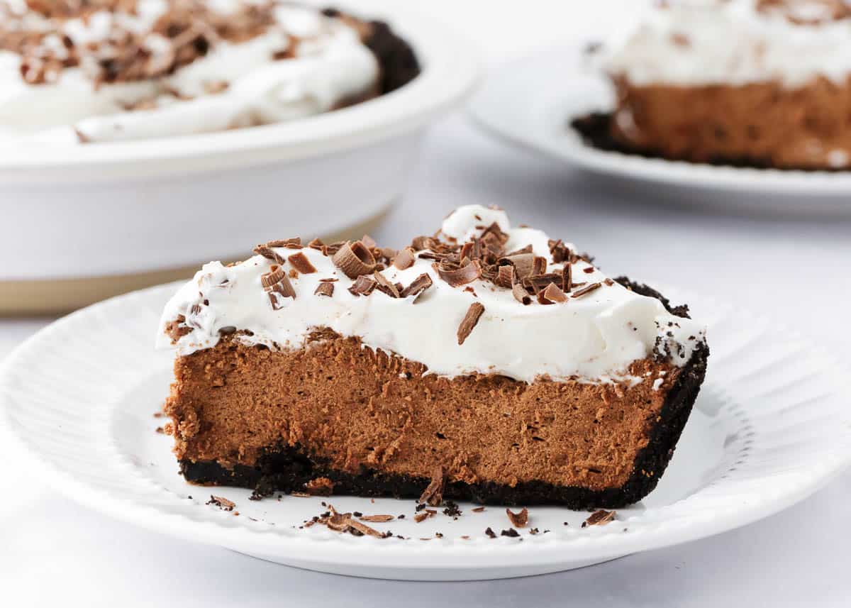 The ultimate chocolate silk pie made with a chocolate crust, silky smooth chocolate filling and topped with homemade whipped cream and chocolate shavings. This dessert is a chocolate lovers dream! A slice of chocolate French silk pie on a plate.