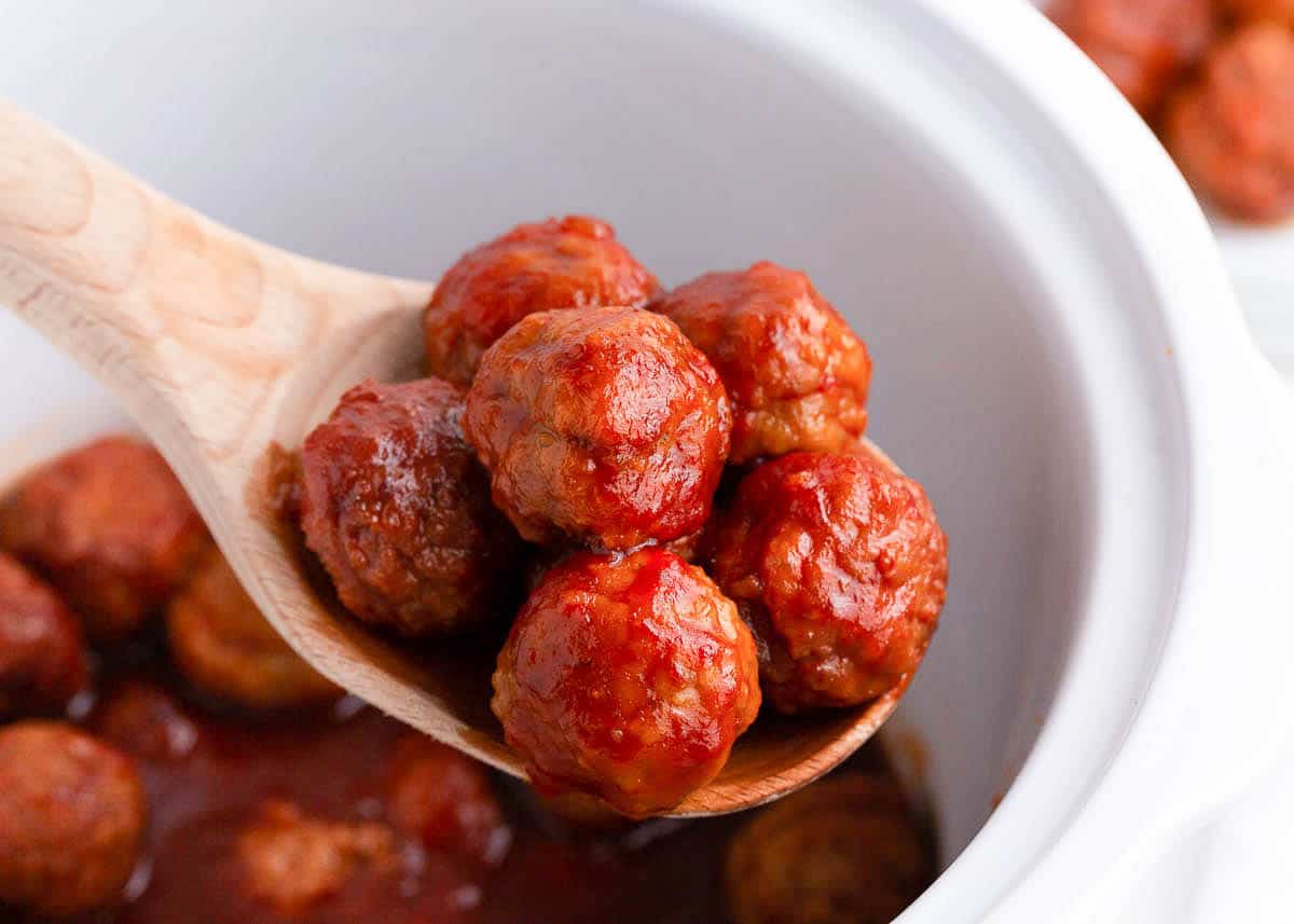 Meatballs on a wooden spoon lifted out of a white crockpot. 