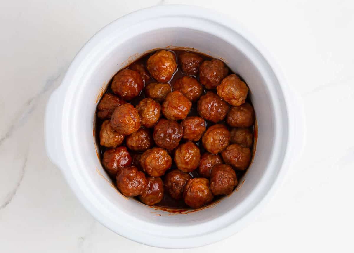 A white slow cooker with cranberry meatballs. 