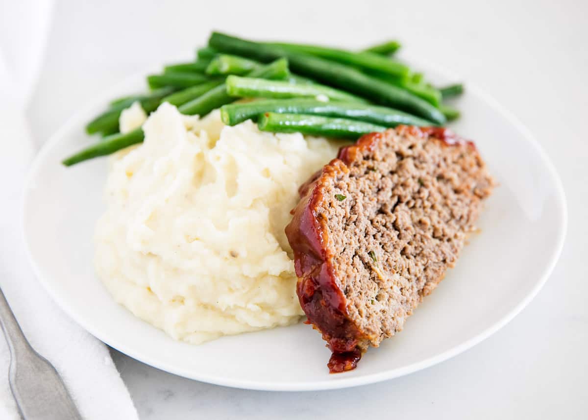Slice of meatloaf on a plate with mashed potatoes and green beans.