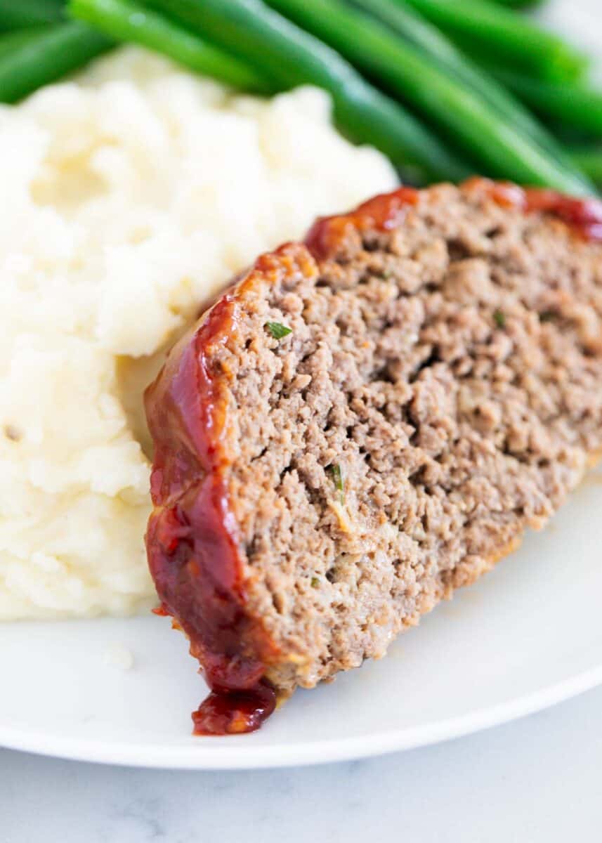 Slice of meatloaf on a white plate with mashed potatoes and green beans.