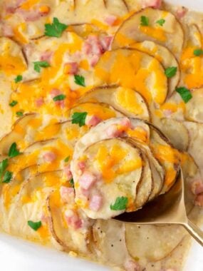 A gold spoon scooping cheesy scalloped potatoes and ham from a baking dish showing the tender potato layers.