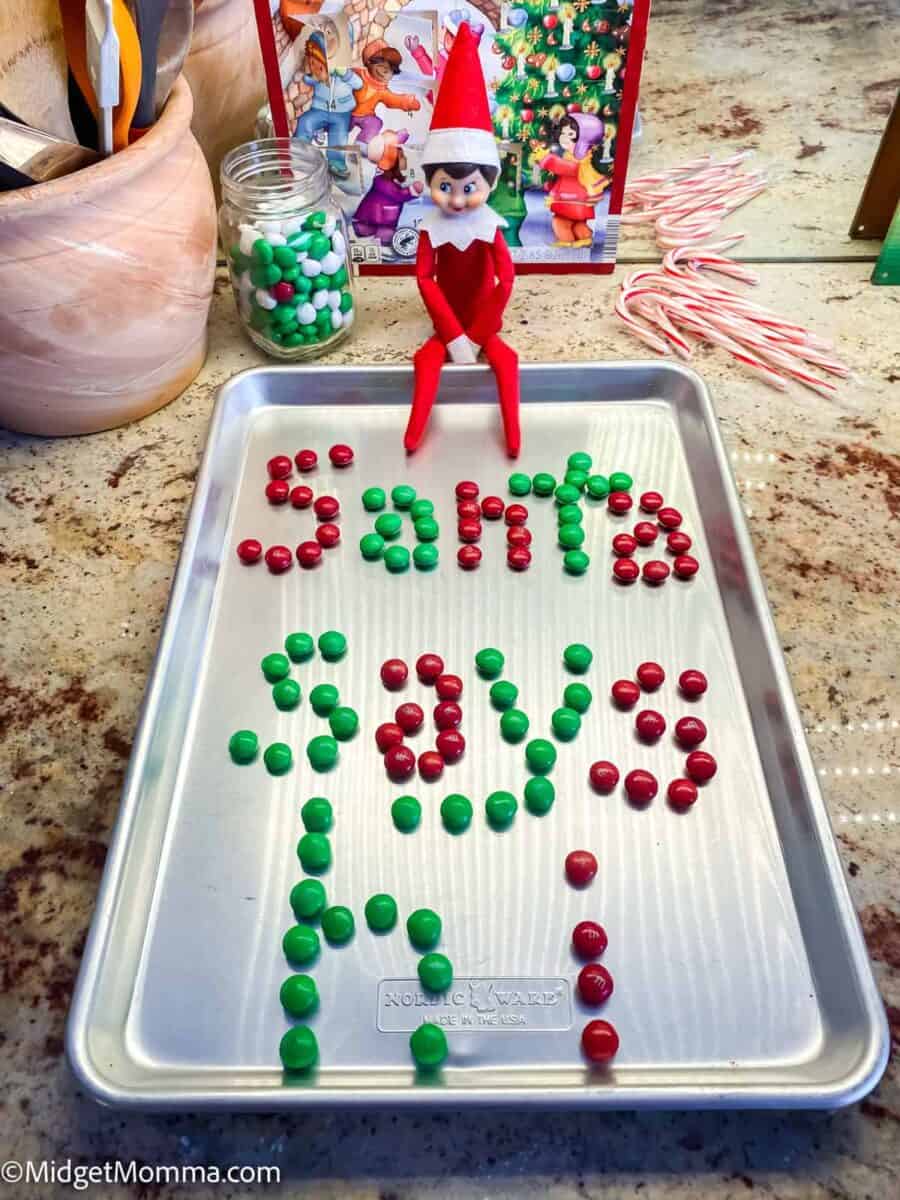 Elf sitting on a baking sheet that says Santa says hi made with m&ms. 