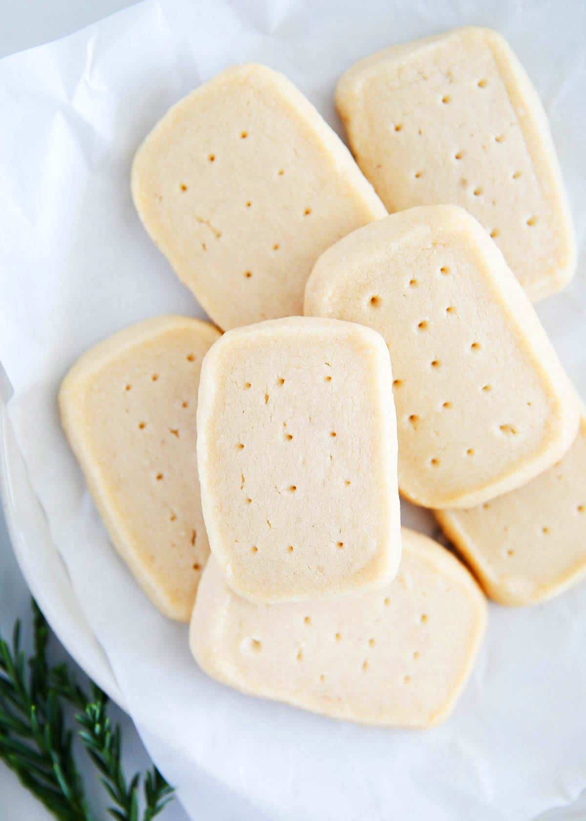 These shortbread cookies are so easy to make with only 5 ingredients, melt in your mouth, and turn out beautiful every time. They are perfect to make cut out, pressed or stamped shortbread cookies. Shortbread cookies on parchment paper on a white plate with a sprig of Christmas greenery.