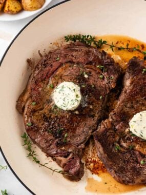 Two oven-baked ribeye steaks in a cast-iron skillet topped with garlic herb butter and fresh thyme sprigs, resting in their pan juices.