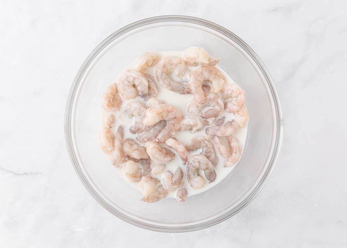 Raw shrimp soaking in buttermilk marinade in a glass bowl.