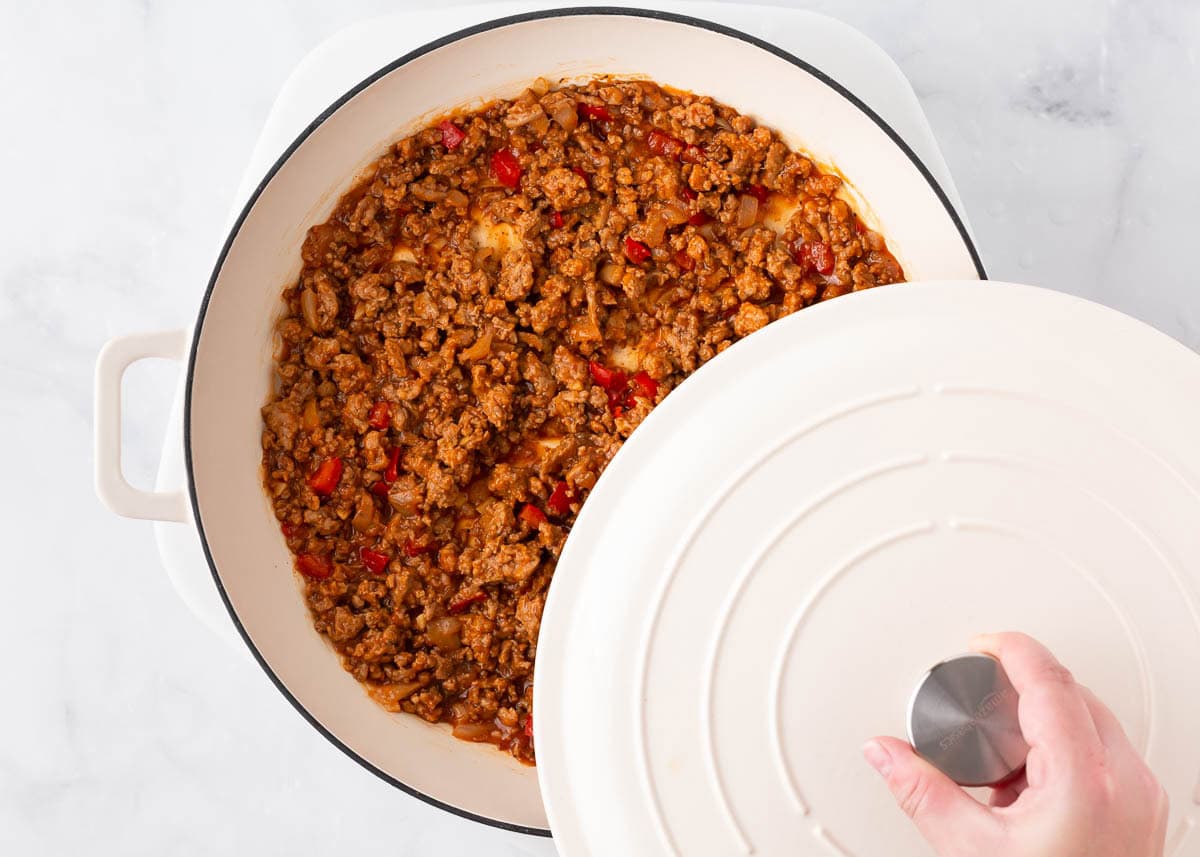 Hand lifting lid off Dutch oven filled with homemade sloppy joes mixture.