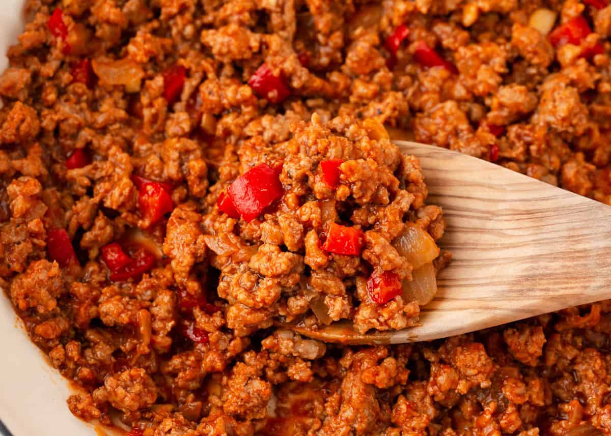 Wooden spoon scooping sloppy joe meat mixture with ground beef, onions, and red peppers.