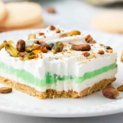 A slice of layered pistachio dessert on a white plate showing creamy filling, shortbread crust and topped with pistachios.