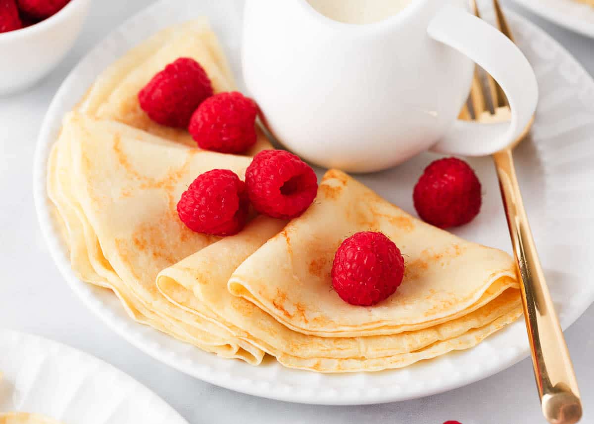 Swedish pancakes on a white plate with raspberries.