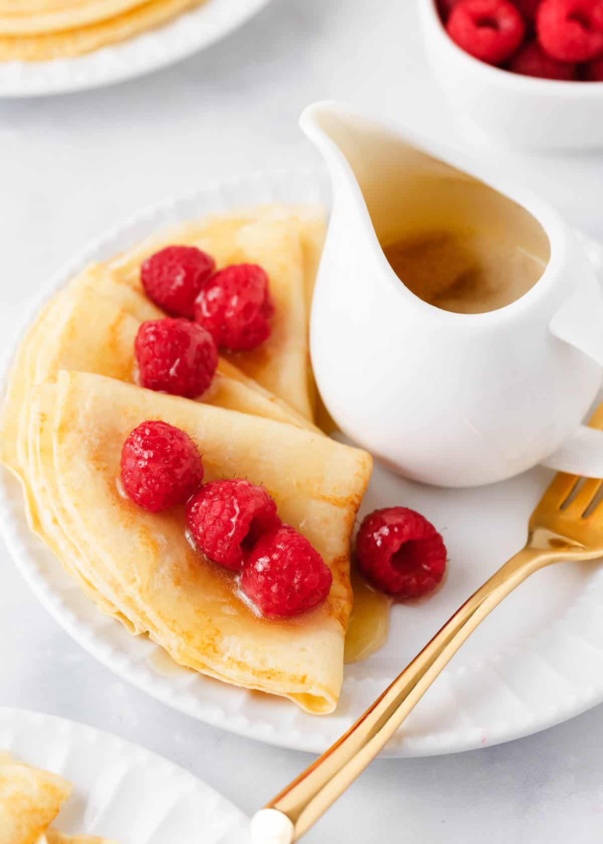 Swedish pancakes with raspberries and syrup on a white plate.