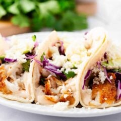 Three baked tilapia fish tacos topped with cabbage and fish taco sauce on a plate.