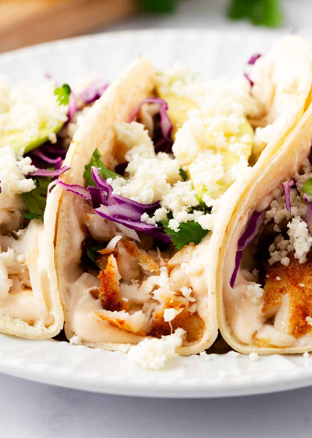 Light and flavorful, this tilapia fish tacos recipe is made with flaky, tender seasoned tilapia and loaded with fresh ingredients. Finished a garlic lime fish sauce, it's a delicious weeknight dinner! Tilapia fish tacos on a white plate.