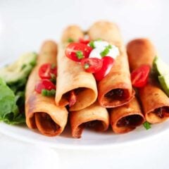 Golden, crispy chicken flautas on a plate with sour cream and tomatoes.