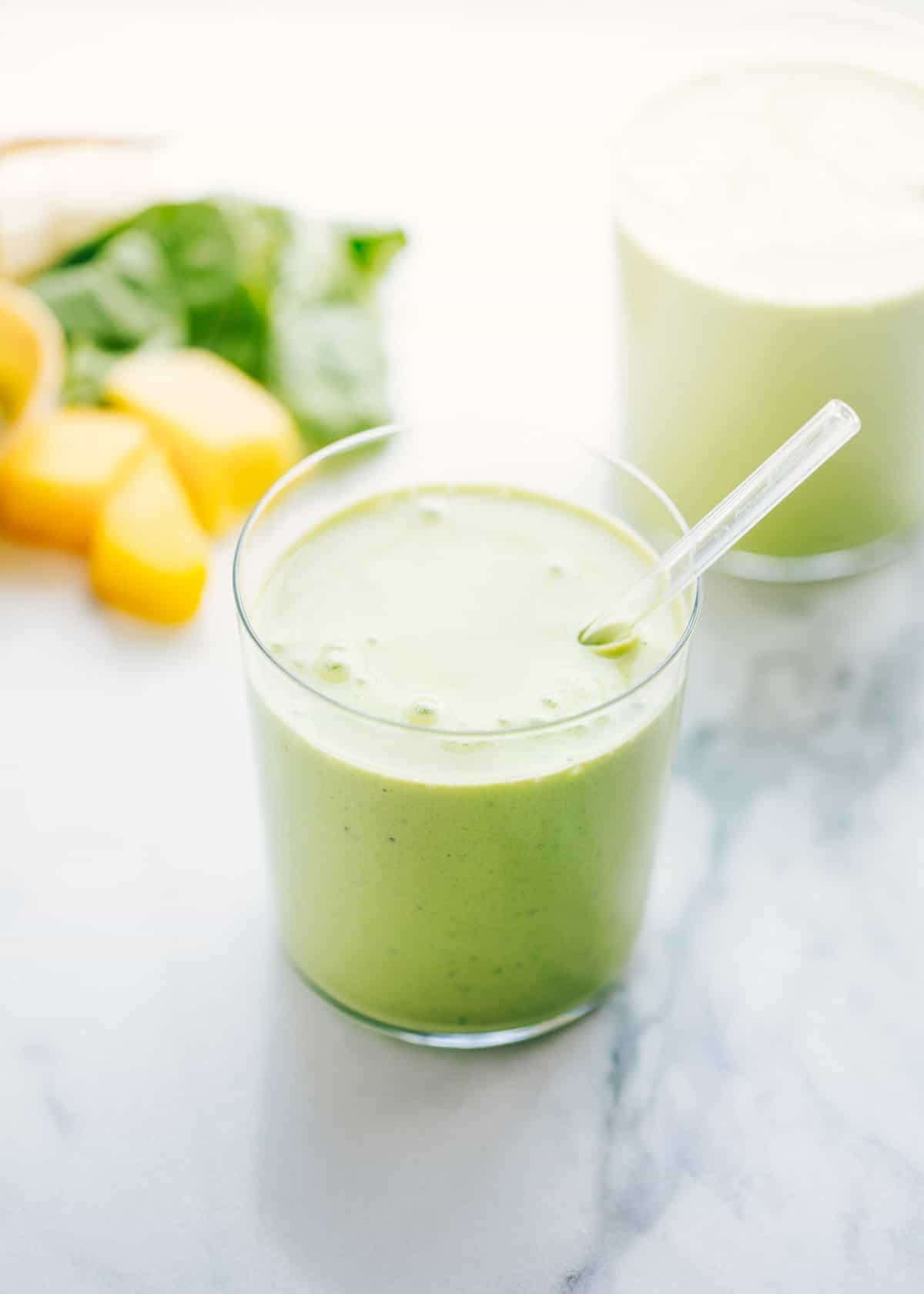 A delicious and healthy green smoothie recipe filled with fruits, greens, milk and yogurt. Perfect for an extra boost of energy and nutrients. A centered glass of green smoothie with a clear plastic straw in it.