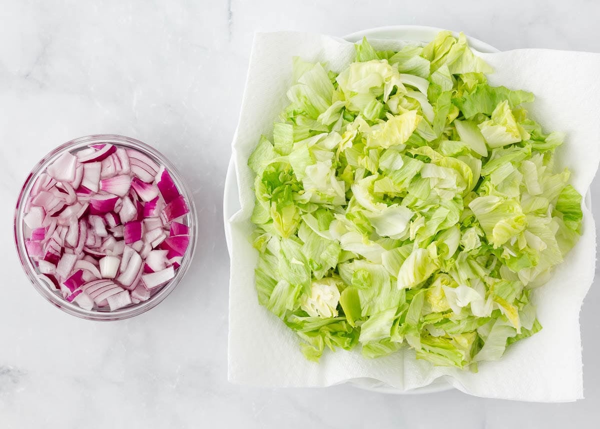 Chopped iceberg lettuce drying on a paper towel-lined plate next to a bowl of diced red onion.