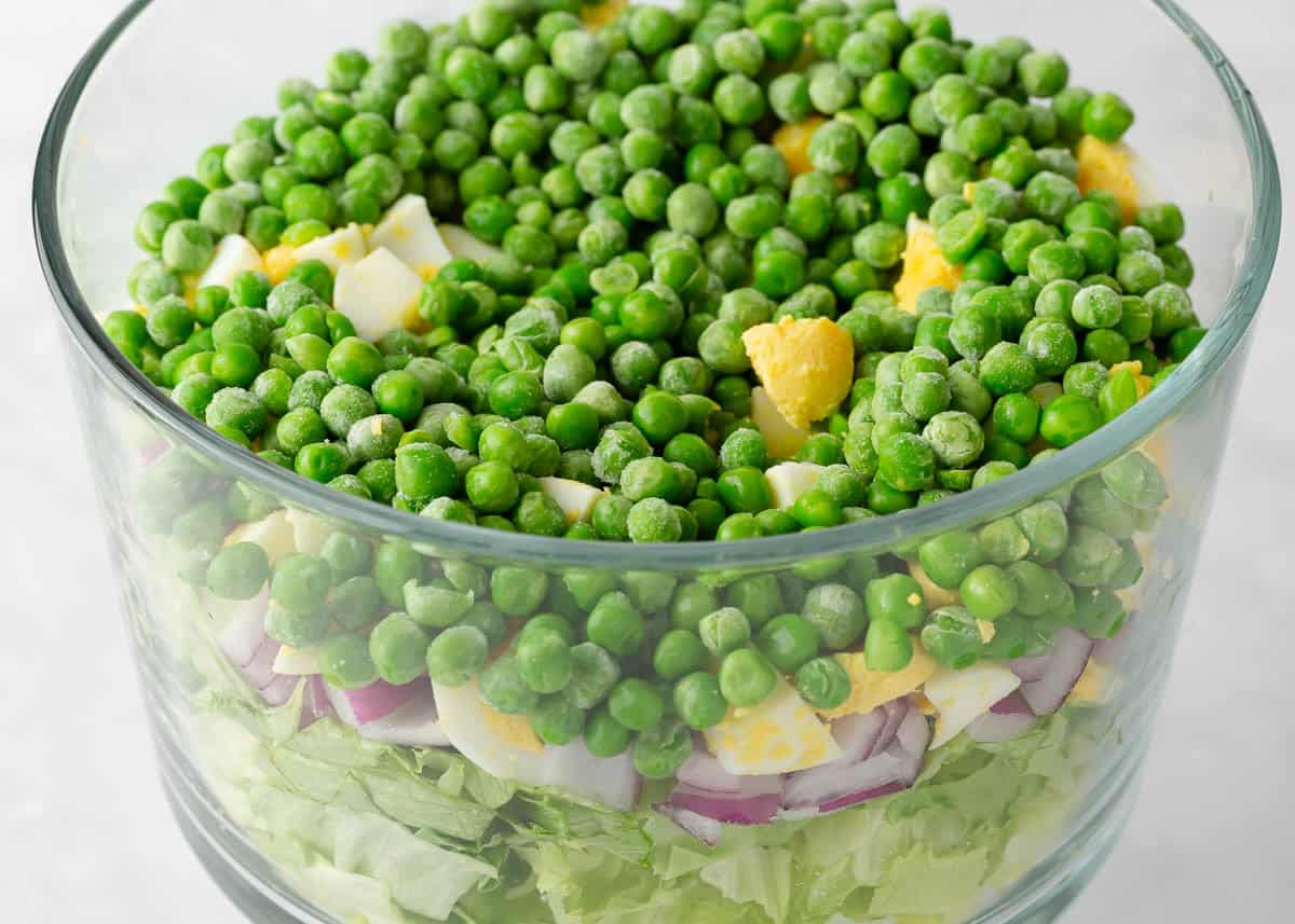 Glass trifle bowl with layers of chopped lettuce, red onion, hard-boiled eggs and green peas for 7 layer salad.