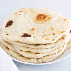 Soft homemade naan bread stacked on a white plate with a white towel nearby.