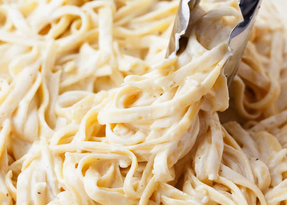 Alfredo sauce with cream cheese tossed with fettuccini being served with tongs.