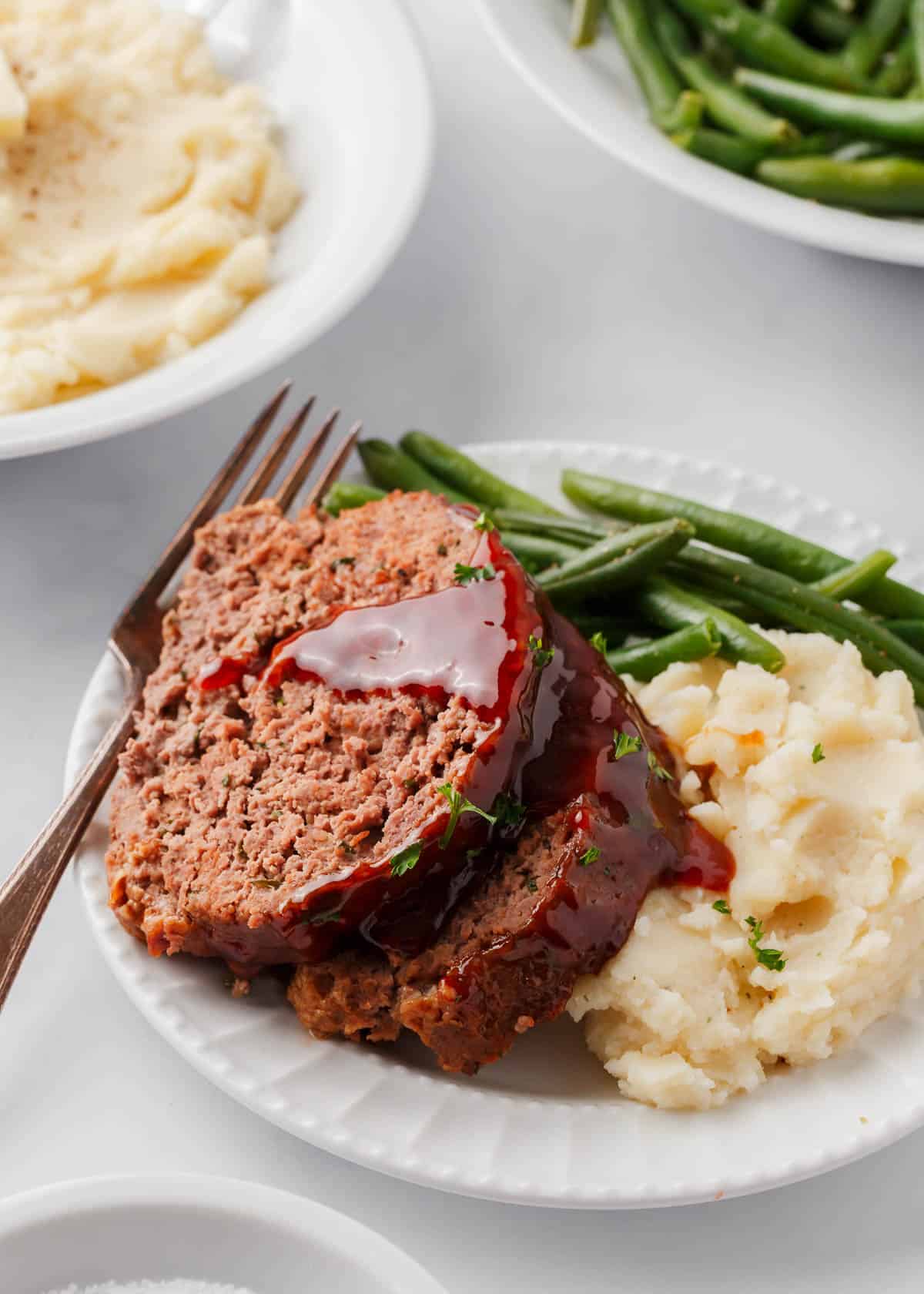 Sliced bbq meatloaf on white plate with mashed potatoes and green beans.
