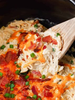 Wooden spoon lifting a scoop of creamy crockpot crack chicken from a slow cooker, topped with melted cheddar cheese, crispy bacon, and chopped green onions.