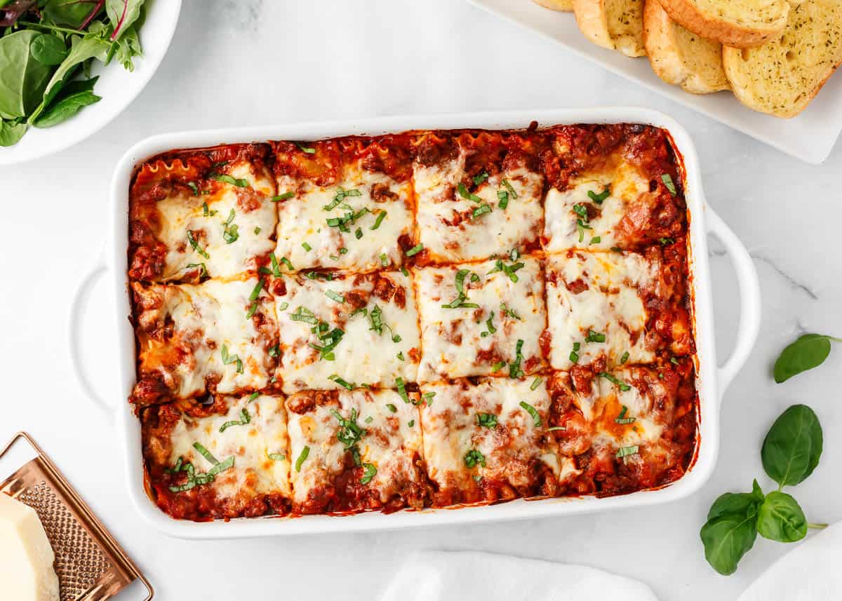 Filled with delicious layers of noodles, cheese and a flavorful beef tomato sauce. This easy lasagna recipe will become an instant hit with your family! Lasagna sliced in white baking dish.