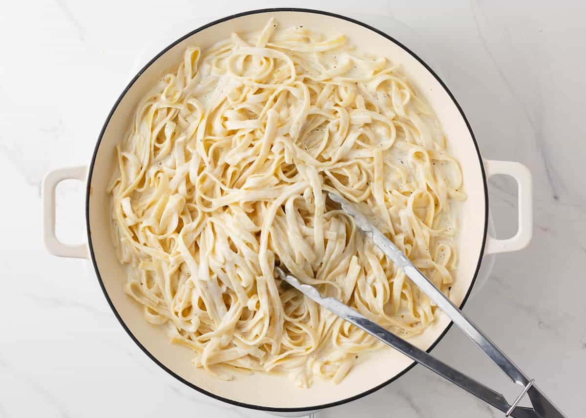 Fettuccine noodles tossed in creamy Alfredo sauce with tongs.