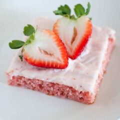 A slice of strawberry sheet cake topped with strawberry cream cheese icing and a fresh strawberry on a serving plate.