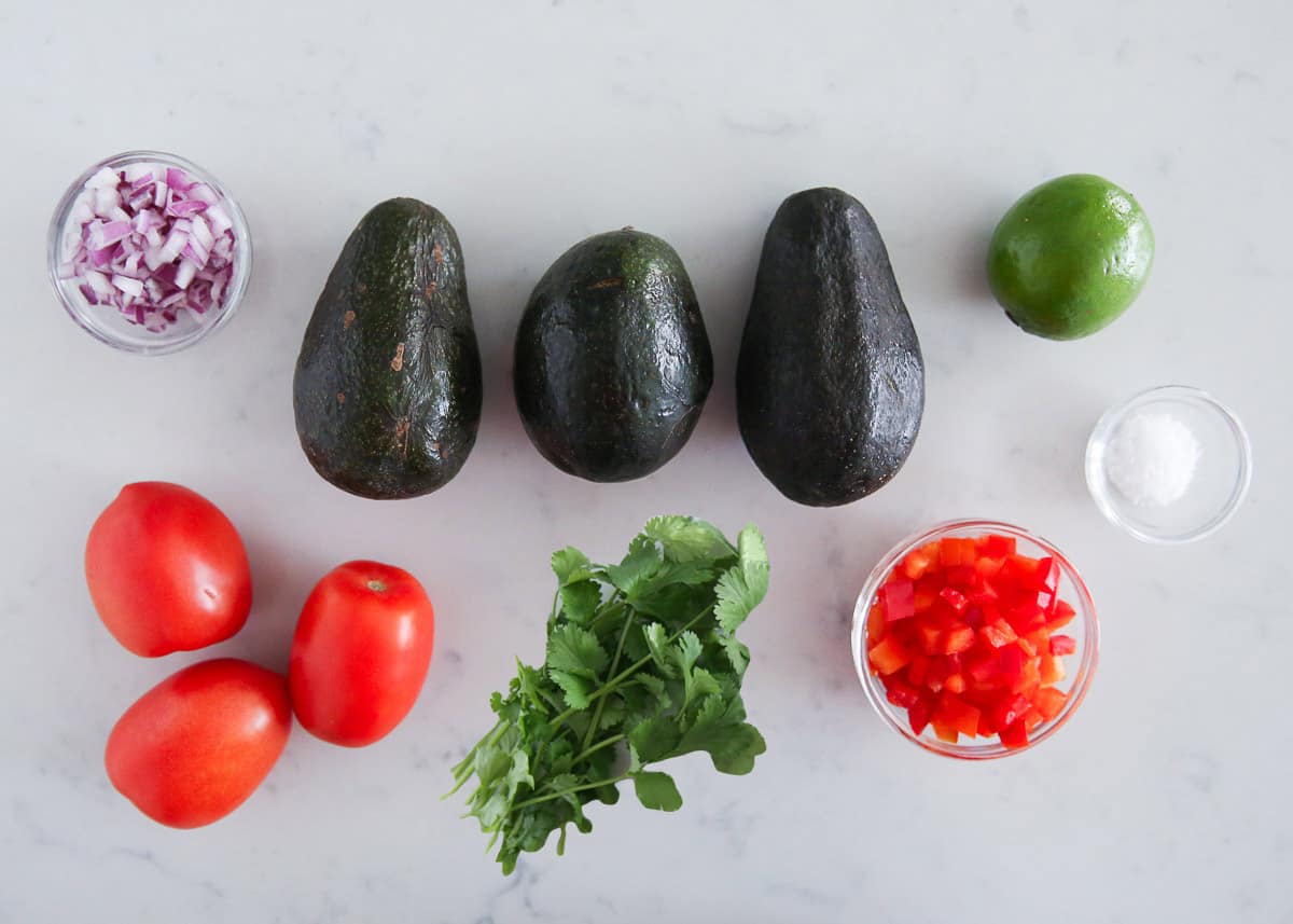 Avocados, tomatoes, red bell pepper, lime, red onion, cilantro and salt on the counter. 