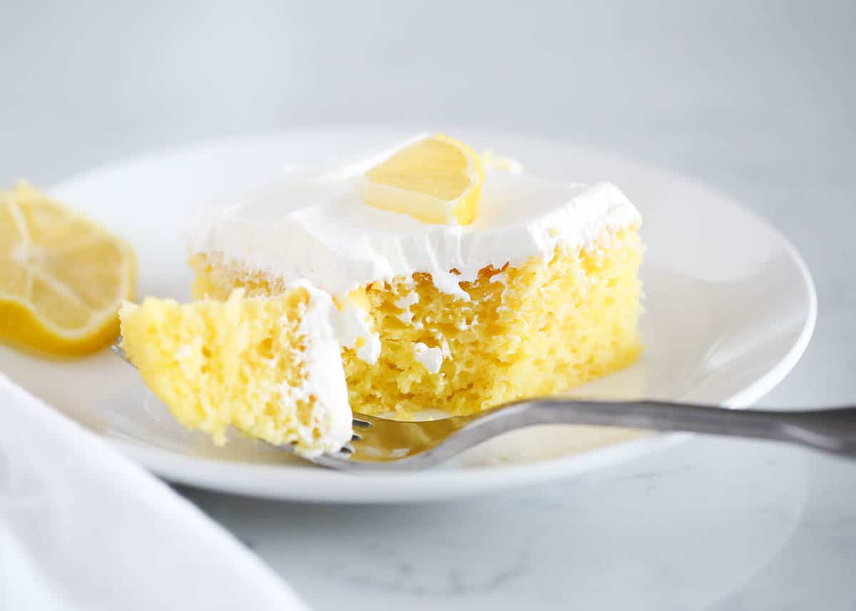 Lemon poke cake on a white plate with a fork taking a bite out of the cake. 