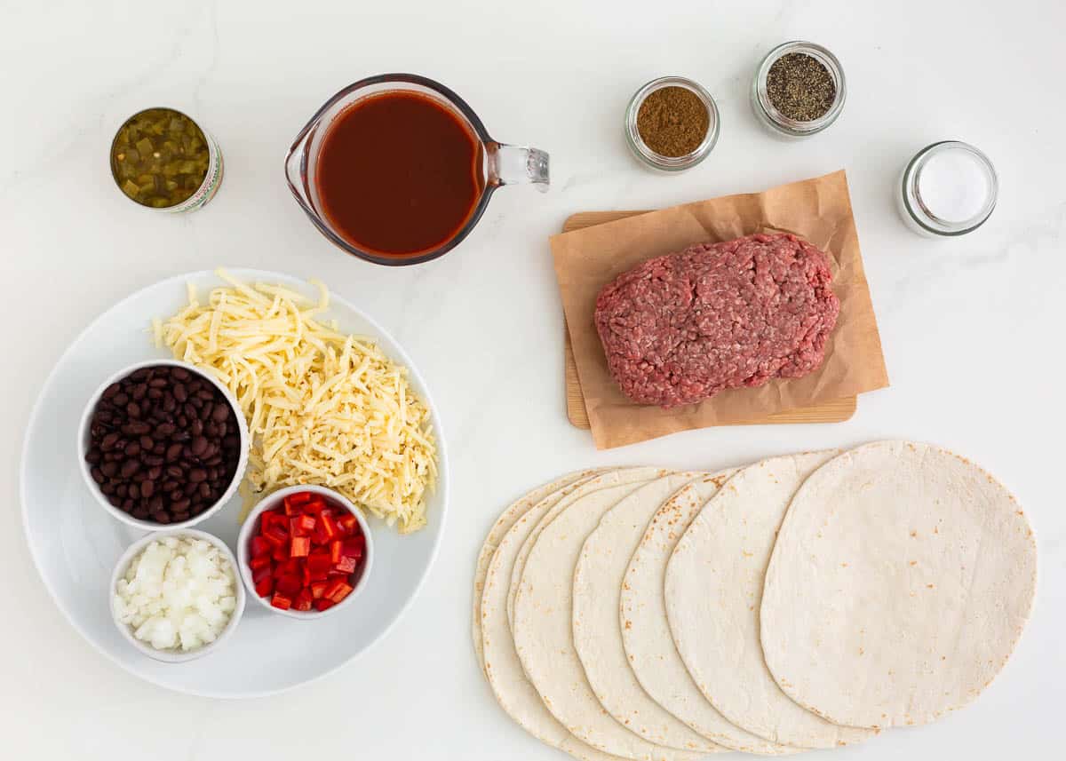 Ground beef, seasonings, red enchilada sauce, beans, flour tortillas and cheese on the counter.