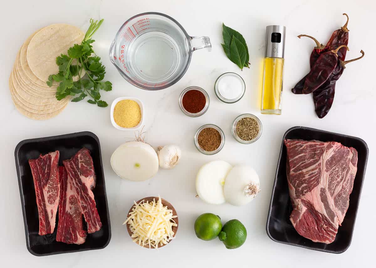 Chuck roast, short ribs, dried chili peppers, onion, garlic, olive, dried herbs and spices, corn tortillas and cheese on the counter. 