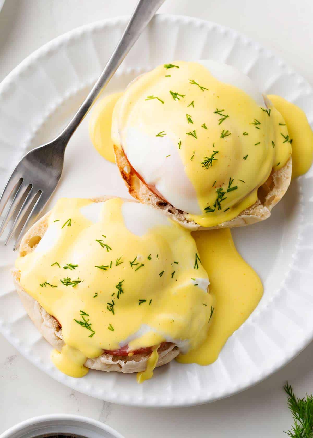 It's easy to make an eggs benedict recipe at home! A toasted English muffin is topped with a slice Canadian bacon and a poached egg then covered with a simple hollandaise sauce. Eggs Benedict on a white plate.