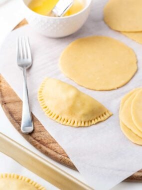 Filled and fork-crimped empanada next to rolled empanada dough discs and egg wash on a wooden board.