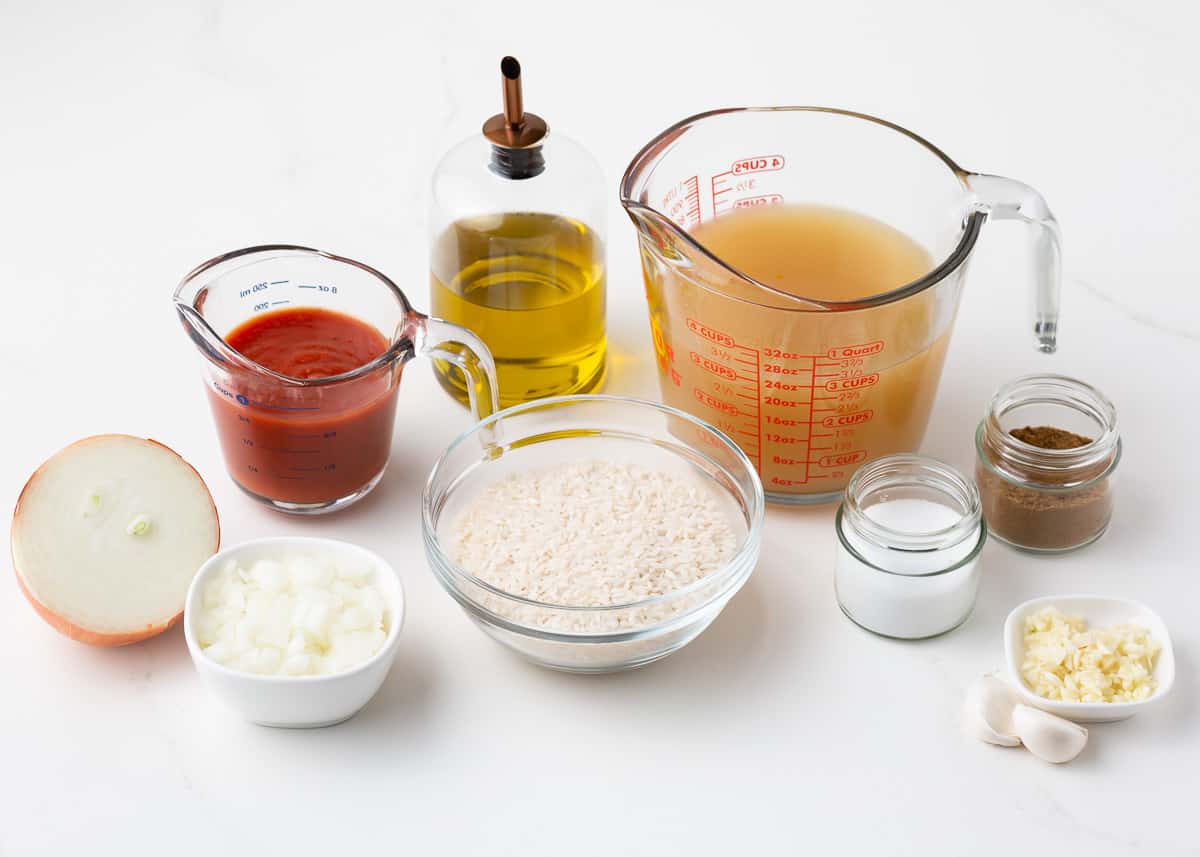 Rice, tomato sauce, chicken broth, onion, garlic, cumin with salt and oil on a counter.