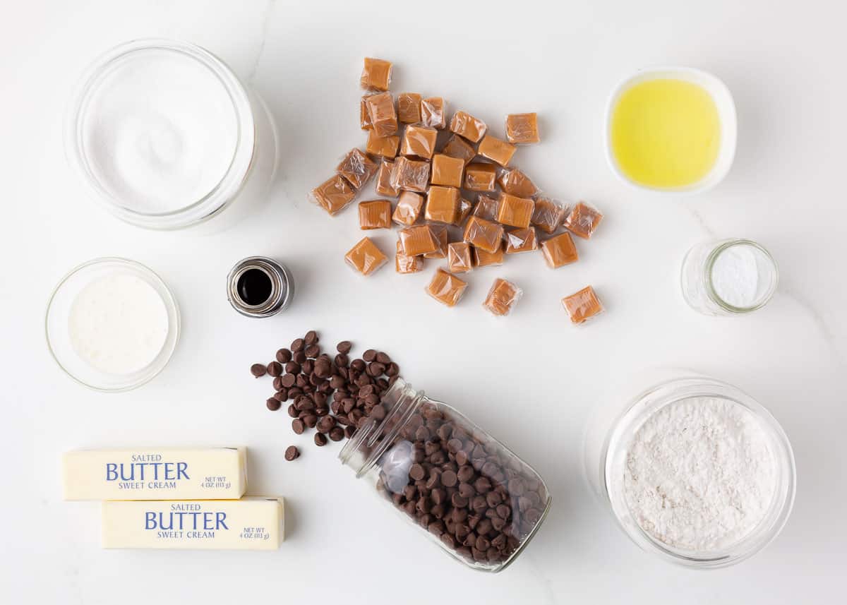 Twix cookie ingredients on the counter including flour, sugar, butter, chocolate chips, egg whites, butter and caramels.