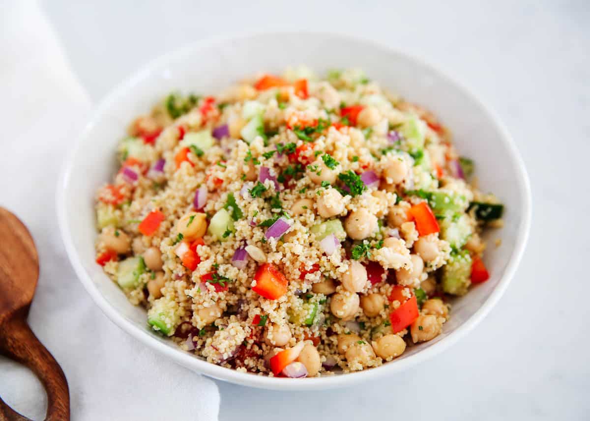 Fluffy couscous, nutty chickpeas, crunchy bell peppers, cucumbers, and sprinkled with briny feta cheese. This couscous salad is tossed in a simple homemade lemon dressing to brighten it up and add some zip to every bite! Couscous salad in a white bowl.