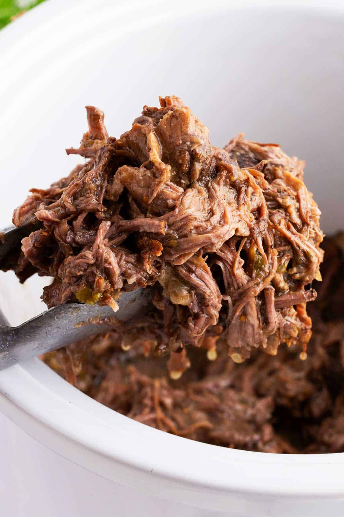 Shredded green chili beef burritos being pulled out of a white slow cooker. 