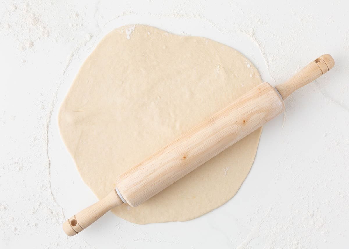 Pizza dough rolled out on a floured surface with a wooden rolling pin.
