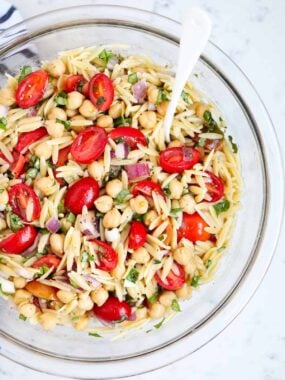 Close-up of orzo salad showing chopped tomatoes, chickpeas, fresh herbs, and red onion in a glass bowl.
