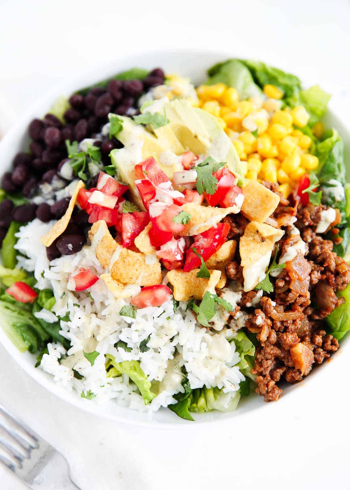 How to make a delicious and easy taco salad recipe that is ready in just 20 minutes! Filled with chopped greens, seasoned ground beef, rice, corn, beans, tomatoes and your favorite salad dressing, it's a flavorful dinner that whole family will love. Taco salad recipe in a bowl.